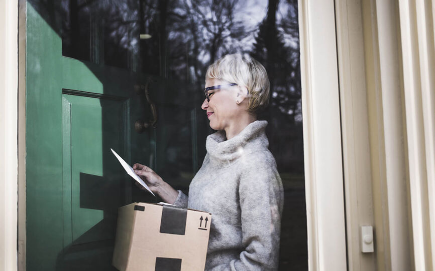 Eldre kvinne har nettopp tatt imot en pakke levert på døren. Esken er i brun kartong med svart pakketeip. Hun smiler mens hun ser ned på et dokument hun holder i hånden.
